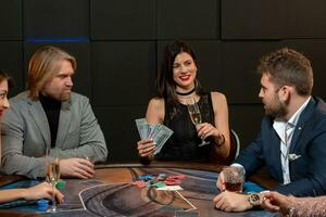 gelukkig donker haar meisje met bankbiljetten en glas van Champagne zittend Bij poker tafel in casino foto