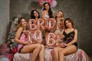Dames in sexy lingerie en bruid in sluier, genieten van een vrijgezellenfeest, zittend Aan bed, Holding ballonnen, lachend. nieuw jaar boom, decoraties. detailopname. foto
