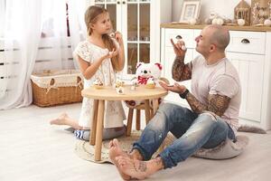 aanbiddelijk dochter vervelend een wit jurk waarmee haar liefhebbend vader. ze zijn drinken thee van een speelgoed- gerechten in een modern kinderen kamer. gelukkig familie. foto