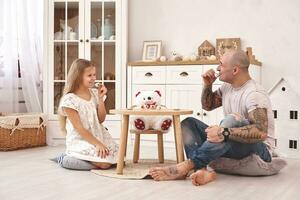 aanbiddelijk dochter vervelend een wit jurk waarmee haar liefhebbend vader. ze zijn drinken thee van een speelgoed- gerechten in een modern kinderen kamer. gelukkig familie. foto