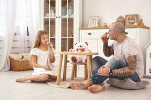 aanbiddelijk dochter vervelend een wit jurk waarmee haar liefhebbend vader. ze zijn drinken thee van een speelgoed- gerechten in een modern kinderen kamer. gelukkig familie. foto