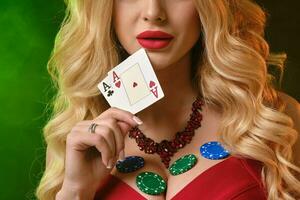 vrouw in rood jurk. tonen twee groen chips en sommige kleurrijk Aan haar borsten, Holding twee azen, poseren Aan kleurrijk rokerig achtergrond. poker, casino foto