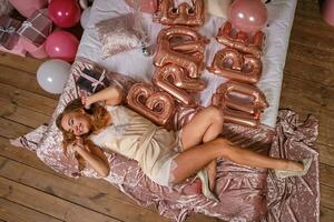 aantrekkelijk dame in sexy beige lingerie is hebben pret gedurende vrijgezellenfeest van bruid, houdende Aan bed, lachend. decoraties, ballonnen. detailopname. top visie. foto