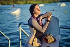 brunette meisje in een grijs coltrui heeft Gesloten haar ogen terwijl poseren met haar wakeboard en staand Aan een pier van de rivieroever. detailopname. foto