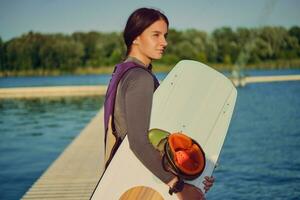 brunette meisje in een grijs coltrui, speciaal sport- hesje en blauw denim shorts is poseren met haar wakeboard Aan een pier van de kust- zone. detailopname. foto