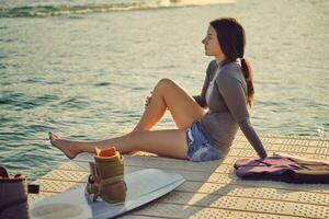 brunette meisje in een grijs coltrui, speciaal sport- hesje en blauw denim shorts is poseren met haar wakeboard Aan een pier van de kust- zone. detailopname. foto