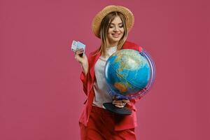 blond meisje in rietje hoed, wit blouse en rood broekpak. ze is lachend, Holding wereldbol, paspoort en ticket, poseren tegen roze achtergrond. detailopname foto