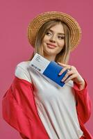 blond vrouw in rietje hoed, wit blouse en rood broekpak. ze lachend, Holding paspoort en ticket terwijl poseren Aan roze studio achtergrond. detailopname foto