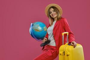 meisje in hoed, wit blouse, rood broekpak. lachend, Holding wereldbol. leunend Aan geel koffer, paspoort en ticket Aan het, poseren Aan roze achtergrond foto