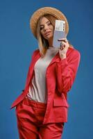 blond dame in rietje hoed, wit blouse en rood broekpak. ze is tonen paspoort en ticket terwijl poseren Aan blauw studio achtergrond. detailopname foto