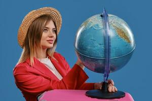 blond model- in rietje hoed, wit blouse en rood jasje. ze is Holding een wereldbol staand Aan roze koffer, poseren tegen blauw achtergrond. detailopname foto