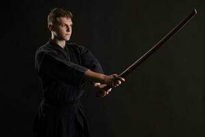 kendo goeroe vervelend in een traditioneel Japans kimono is beoefenen krijgshaftig kunst met de shinai bamboe zwaard tegen een zwart studio achtergrond. foto