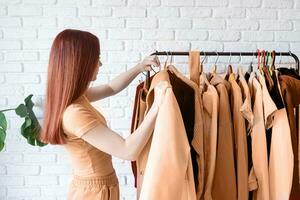 kleur van de jaar 2024, perzik dons. jong mooi vrouw vervelend beige pastel jas, staand in de buurt kleren rek. garderobe verandering foto