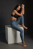 mooi sexy brunette vrouw poseren in studio, zitten, op zoek Bij camera. meisje vervelend modieus jeans en sensueel lingerie. foto