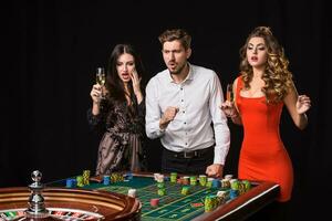 twee jong Dames en Mens achter roulette tafel Aan zwart achtergrond foto