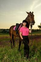 mooi glimlachen meisje jockey staan De volgende naar haar bruin paard vervelend speciaal uniform Aan een lucht en groen veld- achtergrond Aan een zonsondergang. foto