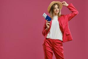 blond vrouw in rietje hoed, wit blouse en rood broekpak. lachend, Holding paspoort en ticket terwijl poseren tegen roze studio achtergrond. detailopname foto