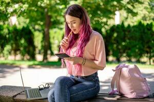 vrouw schrijven in een notitieboekje zittend Aan een houten bank in de park. meisje werken buitenshuis Aan portable computer, kopiëren ruimte. foto