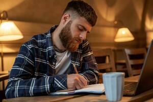 gewoontjes bedrijf Mens of freelancer planning zijn werk Aan notitieboekje, werken Aan laptop computer met slim telefoon, kop van koffie Aan tafel Bij koffie winkel of huis kantoor. foto