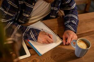 jong gebaard zakenman zit in cafe, huis Bij tafel en schrijft in notitieboekje, in de buurt leugens tablet computer met zwart scherm. Mens is werken, aan het studeren. foto