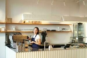 portret van cafe teller met barista meisje werken met koffie machine, maken bestellen voor cliënt in winkel, vervelend blauw schort foto