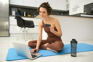 portret van jong vrouw atleet, yoga meisje betalen voor online klassen, inkoop afgelegen opleiding met Sportschool instructeur, zittend Aan haar rubber mat met laptop en credit kaart foto