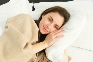 portret van prachtig vrouw aan het liegen in bed, resting Aan hoofdkussen met ogen geopend, op zoek Bij camera en lachend, vervelend pyjama, ontwaken omhoog in ochtend- foto