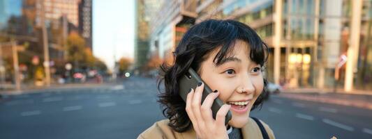 portret van verrast Koreaans meisje, praat Aan mobiel telefoon, hoort verbazingwekkend nieuws over- telefoon gesprek, staat Aan straten van stad met opgewonden gezicht uitdrukking foto