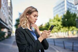 profiel portret van jong verkoopster, zakelijke dame in pak, gebruik makend van mobiel telefoon, lezing Aan smartphone, staand Aan straat buiten kantoor foto