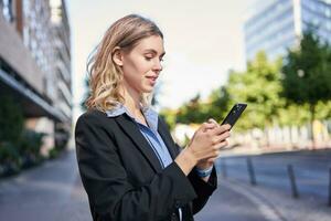 profiel portret van jong verkoopster, zakelijke dame in pak, gebruik makend van mobiel telefoon, lezing Aan smartphone, staand Aan straat buiten kantoor foto