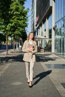 verticaal schot van zakenvrouw wandelen Aan straat met digitaal tablet, gaan naar werk, vervelend beige pak en hoog hakken foto