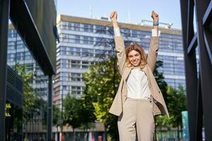 portret van gelukkig jong zakenvrouw optillen handen omhoog, doet vuist pomp, viert zege, wint iets, behaalt doel, staat buitenshuis Aan straat foto