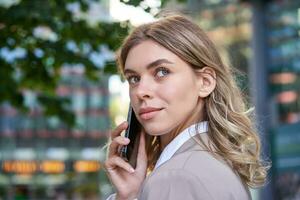 dichtbij omhoog schot van zakenvrouw pratend Aan mobiel telefoon. zakelijke vrouw roeping iemand, op zoek in de omgeving van, staand buitenshuis foto