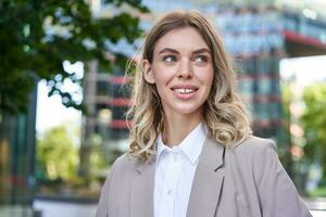 dichtbij omhoog portret van lachend, mooi vrouw 25 jaren oud, vervelend zakelijke kleding, op zoek Vrolijk, staand buiten Aan straat foto