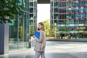 zakelijke vrouw in bedrijf pak, wandelen in stad centrum met werk papieren en mobiel telefoon, roeping iemand foto