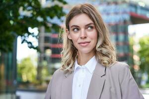 portret van jong geslaagd zakenvrouw in pak, kruis armen Aan borst, glimlach en kijken zelfverzekerd, staand Aan straat van bedrijf centrum foto