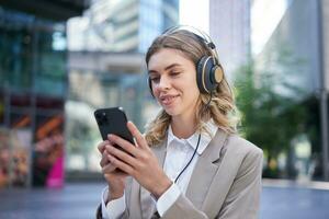 portret van glimlachen jong zakenvrouw luisteren muziek- in hoofdtelefoons en gebruik makend van mobiel telefoon terwijl Aan een bezig stad straat foto