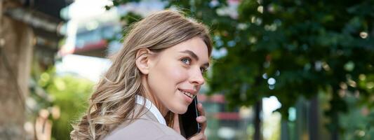 dichtbij omhoog portret van zakelijke vrouw roeping Aan mobiel telefoon, wandelen Aan straat met werk documenten in hand, vervelend bedrijf pak foto