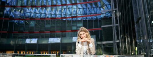 mooi jong vrouw in beige pak, staand in de buurt kantoor gebouwen in stad centrum, glimlachen en op zoek dromerig foto