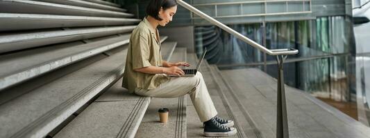 profiel portret van jong Aziatisch vrouw met laptop, meisje leerling zit Aan trap buiten gebouw en types Aan computer, drankjes meenemen koffie foto