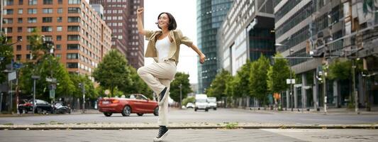 portret van Aziatisch gelukkig meisje jumping en dansen in stad centrum, poseren Aan straten, uitdrukken vreugde en opwinding foto