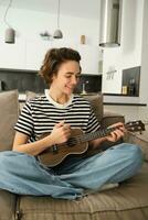verticaal portret van gelukkig jong vrouw spelen ukelele, leert nieuw musical instrument, zit Aan sofa Bij huis, glimlachen met vreugde foto
