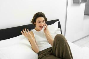 spraakzaam jong vrouw lachend, pratend Aan telefoon, roeping vriend, aan het liegen in bed en hebben gesprek met iemand, resting in slaapkamer foto