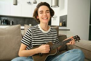 portret van jong modern vrouw, musicus spelen ukulele Bij huis, plukken akkoorden voor nieuw liedje, zittend Aan sofa in leven kamer en glimlachen foto