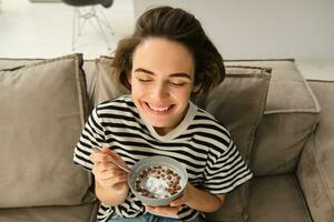 bovenste hoek schot van Vrolijk, schattig jong vrouw Aan bank, aan het eten kom van granen met melk en lachend, genieten van haar ontbijt foto