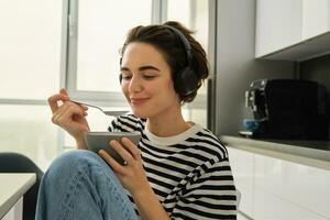 dichtbij omhoog portret van lachend, mooi jong vrouw in koptelefoon, aan het eten granen voor ontbijt en luisteren muziek- of e-boek, Holding lepel met kom foto