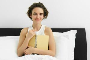 portret van vrouw zittend in bed met logboek, schrijven in haar dagboek, gedachten of gevoelens in de ochtend, Holding notitieboekje en glimlachen foto