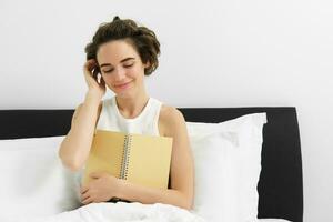 portret van mooi vrouw in bed, zittend met haar logboek, schrijven in dagboek en lachend, instoppen haar- achter oor, lezing notitieboekje foto