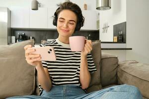 glimlachen meisje aan het kijken TV tonen Aan smartphone, vervelend koptelefoon, drinken thee en zittend Aan sofa foto