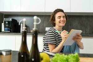 dichtbij omhoog van Vrolijk, glimlachen jong vrouw schrijven in notitieboekje, creëren recept, lijst van maaltijden voor deze week, zittend in de buurt groenten en olijf- olie, Koken in de keuken foto